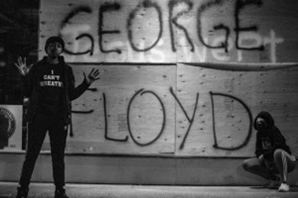 A picture of a young black man standing on a sidewalk with his hands in the air. The wall in the background has the words "George Floyd" spray painted on it.