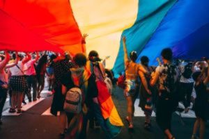 Many people marching together under a large rainbow banner.