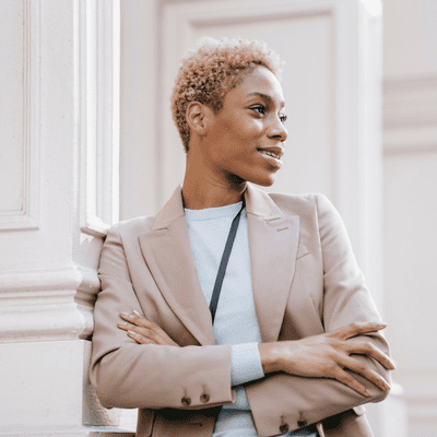 A woman in business attire is smiling and waiting while leaning against a pole.